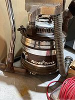 Filter Queen canister vacuum cleaner showing polished metal body with logo, hose, and attachments on concrete floor.