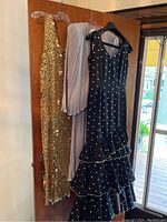 Three dresses on hangers including a gold-tone chain metal slip dress, a light gray bejeweled jacket, and a black flamenco dress with white polka dots and ruffles.