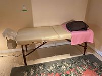 Photo of beige folding massage table set up against a wall on a decorative rug, with blanket and storage bag on top.