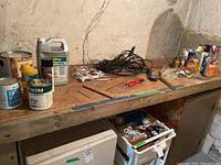 Wooden tabletop holding various cans, bottles, tools, and electrical cords. Items include cleaning solutions and a variety of tools.