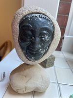 Front view showing the detailed black carved stone face with textured hood stone and smooth stone base on tiled surface.