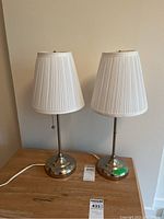 Pair of matching table lamps shown side by side on a wood surface against a beige wall. White pleated fabric shades with metal stems and round bases are visible.