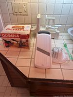Olympia pasta machine with original box, OXO mandolin slicer, and spiralizer on tiled countertop.