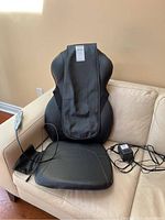 Full view of the black Homedics massage chair pad showing the padded backrest and seat area, with attached power adapter and wired remote control.