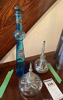 Photo showing two clear glass oil lamps with detailed metalwork and one tall blue glass bottle with stopper and metal accents.
