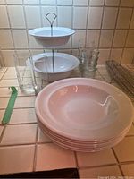 Photo showing six stacked white ceramic pasta bowls alongside a white two-tier bowl stand, one tall glass, four short glasses, and a metal wire basket on a tiled kitchen counter.