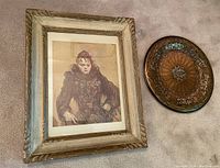 Framed print of a human figure alongside decorative copper plate on carpeted floor.