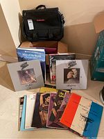 Vinyl records, books, and black Barclay branded laptop bag arranged on floor with some box sets visible