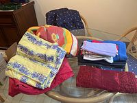 Photo showing a collection of various table linens including placemats, tablecloths, runners, and folded napkins on a glass table.