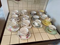 Overview photo showing 14 vintage teacups with matching saucers on tiled countertop. Various floral patterns and colors including white, yellow, pink, blue, purple with gold trim accents. One yellow cup with gold handle and rim is prominent. Cups are arranged with saucers beneath.