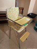 Front angle view of vintage doll high chair showing floral vinyl seat and backrest, yellow tray with warning label, and chrome metal frame.