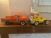 Two vintage Tonka trucks side by side on wooden shelf: orange CAA service truck and yellow box truck