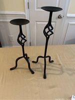 Two black metal candle holders with varying heights standing on a beige surface, showing front and side views.