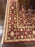Close-up of rug corner showing detailed floral border design, red background with gold and green accents, and visible fringe.
