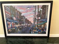Framed poster showing a vibrant Italian Market street scene with numerous vendors and lights overhead, viewed from the street level, image taken from an angle showing the full frame and poster in good condition.