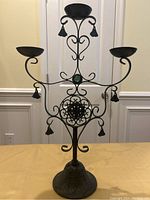 Front view of decorative black wrought iron candle holder with three candle cups and tassels, ornate scrollwork, and green accent on round base.