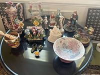 View of eleven ceramic items on black table including figurines, a decorated bowl, floral vases, a mortar and pestle, and other decorative pieces.