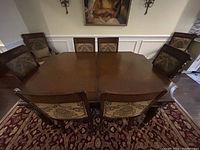 Photo of rectangular wood dining table surrounded by eight wood chairs with floral tapestry upholstery, on a patterned rug against cream walls.