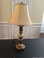 Photo showing a decorative table lamp with a beige fabric bell-shaped shade and an ornamental base with gold and cream tones.