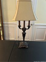 Full view of the decorative table lamp showing the beige fabric shade and ornate bronze/black base with detailed motifs and candle-style accents.