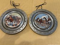 Two pewter-rimmed wall plates with western ceramic center artwork side by side on yellow cloth background.