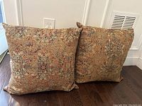 Pair of large square pillows with rust, blue, and tan intricate patterns photographed on a wooden floor near a power outlet and vent.