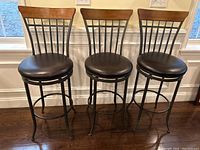 Three dark brown wooden bar stools with black cushioned seats arranged side by side showing full front view.