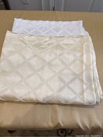 Two folded tablecloths displayed, one white and one beige with diamond jacquard pattern