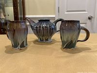 Front view of ceramic tea pot flanked by two cups on a wood surface under indoor lighting.