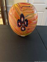 Front view of the oval-shaped colorful abstract glass vase showing swirling orange and yellow colors with blue oval spots on a black surface.