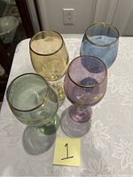 Photo showing four colored long stem wine glasses with gold rims on a white tablecloth, positioned closely together from top view.