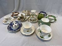Overview of all 12 bone china cup and saucer pairs arranged in two staggered rows with a green apple for scale.
