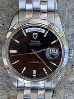 Front close-up of black baton dial showing day display at 12 o’clock and date magnifier at 3 o’clock.