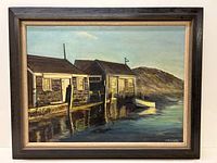 Framed oil on canvas painting showing full lakeside scene with wooden boathouses, boat, and distant hill.