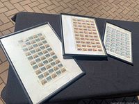 Three framed sheets of vintage stamps shown resting on a black surface outdoors, all frames are black with white mat board and display rows of identical stamps.