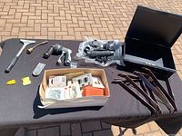 Overview of black metal box and assorted tools including pry bars, hose nozzle, scraper, and small parts box placed on black cloth outdoors