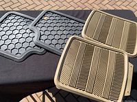 Two sets of car floor mats: one black pair with hexagonal pattern and one beige pair with linear grooves, laid out on a black fabric surface.