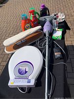 Full view of all cleaning and laundry items displayed on a small black table outdoors on a brick surface. Items include bottles of Tide detergent, fabric steamer, ironing board, glove box, and other cleaning products.