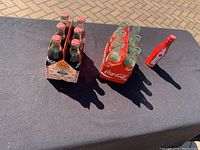 Seven Coca-Cola collectible bottles displayed: six empty in cardboard holder with basketball and Trail Blazers graphics, one solid red unopened Big Ben bottle separate.