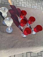 Five red glasses and two clear crystal candlesticks displayed on a brown cloth outdoors showing some shadows.