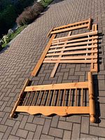 Various pine wood parts of an older double bed frame laid out on a paved surface. Pieces include a slatted base, side rails, and headboard components with visible wear and damage.