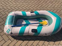 Full view of the inflated white and teal inflatable boat with a blue paddle lying across it, displayed on an outdoor paved surface.