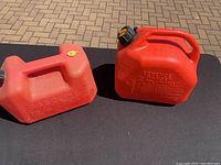 Two red plastic fuel containers with handles and spouts placed on a black surface outdoors on paved ground.