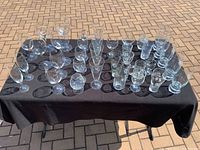 Overview of 34 various clear glass drinkware pieces arranged on black tablecloth outdoors on brick patio.