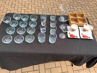 Photo showing all glassware items arranged on black tablecloth including glass bowls, dessert cups, small glasses, wine glasses, porcelain cups and saucers, and wooden organizer.