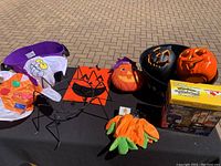 Photo showing Halloween decorations laid out on a black cloth on pavement: two plastic pumpkins (one orange, one black), two pumpkin face fabric decorations on metal stands, and an orange-green cloth corn decoration.