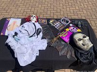 Overview of Halloween masks and decorations laid on black cloth including ghost figure with mask, large skull mask, bat wings, spider prop, DVDs, and other Halloween items.