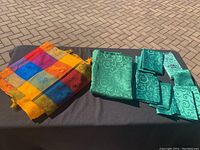 Green tablecloth folded with twelve matching green napkins displayed alongside bright multicolor patchwork tablecloth on black surface outdoors on paving stone background.