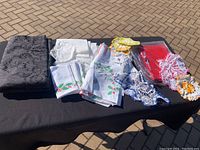Top view of assorted textile items including a large dark grey patterned tablecloth, folded white and printed cloth napkins, crocheted doilies in white and orange, and red textiles with lace detailing.