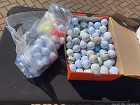 Box filled with approximately 70 worn golf balls including some Titleist and other brands, all mostly white with visible marks and dirt. A plastic bag next to the box also contains additional golf balls.